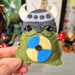 May include: A green felt creature ornament featuring a grey Viking helmet with white horns and a round shield divided into blue and yellow quadrants with a brown center. The figure has black eyes and is stitched with light green thread along the edges.