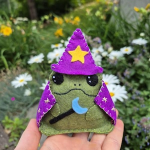 May include: A green felt frog plush toy wearing a purple wizard hat with a yellow star and a matching purple cape featuring embroidered gold stars. The frog holds a small black wand topped with a light blue crescent moon shape.