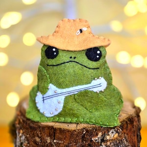 May include: A green felt frog plush toy wearing a tan hat with a small white insect detail. The frog has large black eyes and is holding a white felt banjo with silver bead accents. The stitching is visible along the edges of the felt.