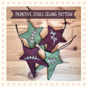 Puede incluir: Cuatro adornos de fieltro en forma de estrella con diseños botánicos bordados en verde y burdeos oscuro. Cada estrella tiene un cordón negro para colgar y puntadas de festón visibles en los bordes. El texto PRIMITIVE STARS SEWING PATTERN aparece.