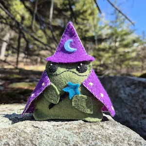 May include: A handcrafted felt frog, styled as a wizard. The green frog has black eyes and wears a purple hat and cape. The hat features a blue crescent moon, and the cape is adorned with yellow stars. It holds a blue star-shaped wand.