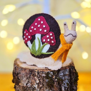 May include: A felt snail figurine with a beige body and a dark brown shell. The shell features three red mushrooms with white spots and green grass appliqués. The snail wears a bright orange scarf around its neck and has two small eyes.