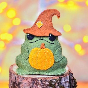Puede incluir: Un peluche de rana de fieltro verde que lleva un sombrero puntiagudo naranja con un parche cuadrado cosido en amarillo. La rana tiene ojos negros y boca cosida, y sostiene una calabaza de fieltro naranja brillante con líneas verticales.