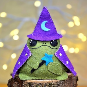 May include: A green felt frog figurine wearing a purple felt wizard hat with a light blue crescent moon and a matching purple cape featuring small yellow star embroidery. The frog holds a small light blue felt star wand in its front paws.