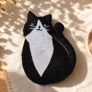 May include: A black and white felt cat-shaped coaster. The cat has a white chest and face with black ears and a tail. The coaster is sitting on a beige surface.