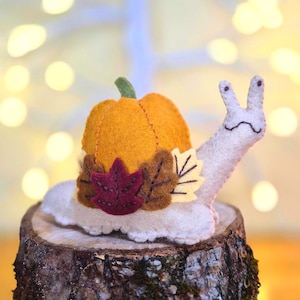 May include: A felt snail figurine with a cream-coloured body and a bright orange pumpkin shell. The pumpkin is decorated with three small felt leaves in shades of deep red, brown, and pale yellow, featuring visible dark brown stitched details.