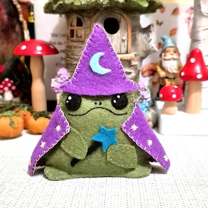 Puede incluir: Un peluche de rana de fieltro verde con un sombrero de mago morado con una luna creciente azul claro y una capa morada a juego con pequeños bordados de estrellas amarillas. La rana tiene ojos negros cosidos y sostiene una varita de estrella.