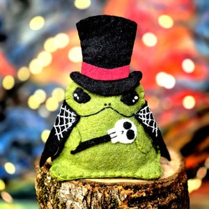 May include: A green felt frog plushie wearing a black top hat with a red band. The frog has black cape-like wings with white spiderweb embroidery and holds a small white skull in its hand. The plush is crafted with visible stitching.