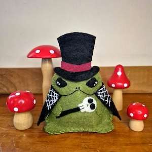 May include: A green felt creature wearing a black top hat with a red band and a black cape with white spiderweb embroidery. The creature holds a small white skull and is surrounded by four red and white wooden mushroom-shaped decorative items.