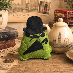 Puede incluir: Un peluche de fieltro verde lima con forma de criatura redondeada, que lleva un sombrero bombín de fieltro negro y un bigote de fieltro negro. El peluche tiene dos pequeños ojos negros con reflejos blancos y sostiene un paraguas negro.