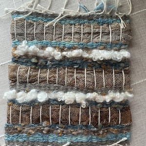 May include: Handwoven textile art piece with a rectangular shape. The piece features a combination of brown, blue, and white yarns, creating a textured surface. The design incorporates woven sections and fluffy, cloud-like elements. The edges have fringe.