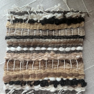 May include: Handwoven textile art piece with a rectangular shape. The piece features horizontal bands of varying textures and colors, including white, beige, brown, and black. The top and bottom edges have loose, white fringe.