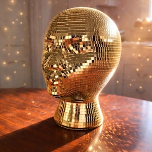 Puede incluir: Una escultura de cabeza dorada con diseño de mosaico de bola de discoteca. La cabeza está montada sobre una base de pedestal, reflejando la luz. La escultura está sobre una mesa de madera oscura, con una luz suave y cálida al fondo.