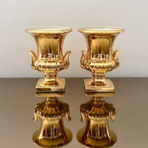 May include: Two matching metallic gold urn-shaped vases with a reflective finish. Each vase features a wide flared rim, two decorative side handles, and a square base. The interior of the vases is white, and the exterior has a smooth, polished surface.