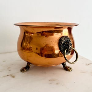 May include: A polished copper bowl with a rounded body, featuring three ornate claw-shaped feet and a decorative lion head handle on the side with a circular ring. The metallic surface has a warm, reflective orange-brown tone and smooth finish.