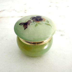 May include: A small, round, light green stone trinket box with a polished finish. The lid features a hand-painted floral design with dark purple and brown pansy-like flowers and green leaves. A thin, shiny gold-tone metal band encircles the centre.