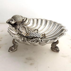May include: A silver-toned metal dish shaped like a scalloped shell, featuring three ornate feet designed as sea creatures. Inside the bowl rests a metallic link bracelet with alternating silver and gold-coloured segments, creating a decorative display.