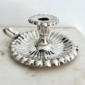 May include: A silver candlestick holder with a fluted base and a handle. The candle holder features a flower-shaped top and a central cup for a candle. The design is detailed, with a reflective surface. The base is approximately 15 cm in diameter.