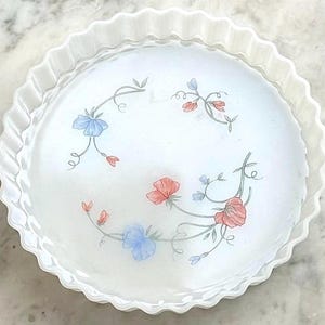May include: White, round pie dish with a scalloped edge. The dish has a floral design with blue and red flowers and green stems. The pie dish is made of a translucent material.