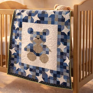 Puede incluir: Una cuna de madera con un edredón de patchwork azul y blanco con un diseño de oso de peluche. El edredón tiene un borde de cuadrados azules y estrellas blancas. Se ve un oso de peluche en la cuna.