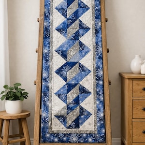 Könnte beinhalten: Ein blau-weiß gesteppter Tischläufer mit geometrischem Design, der auf einer Holzleiter ausgestellt ist. Der Läufer zeigt Rauten in Blau- und Grautönen mit einem Rand aus Schneeflocken. Die Leiter ist aus hellbraunem Holz.