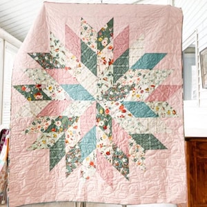 May include: A handmade quilt with a starburst design. The quilt features a light pink background with a central star pattern composed of floral and geometric shapes in shades of pink, green, and blue. The quilt is hanging, showcasing its intricate stitching.