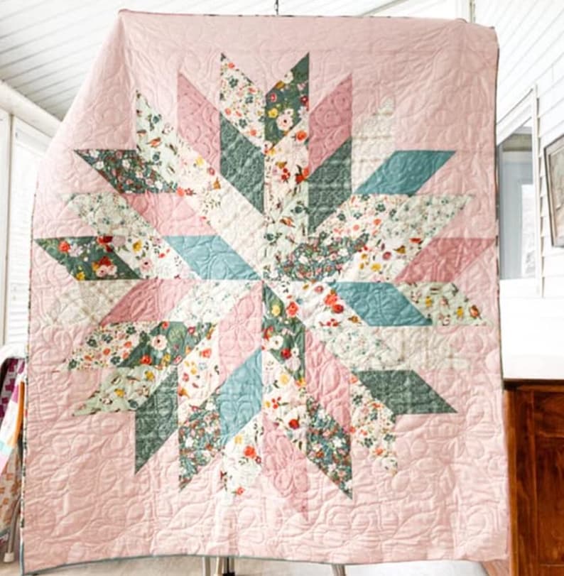 May include: A handmade quilt with a starburst design. The quilt features a light pink background with a central star pattern composed of floral and geometric shapes in shades of pink, green, and blue. The quilt is hanging, showcasing its intricate stitching.
