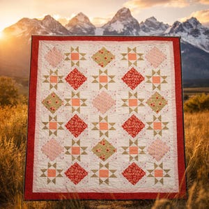 Pode incluir: Um edredom feito à mão com uma borda vermelha e um fundo branco. O edredom apresenta um padrão repetitivo de formas de diamante vermelhas, rosa e verdes, juntamente com detalhes de estrelas e quadrados dourados. O edredom é exibido ao ar livre com um cenário de montanha.