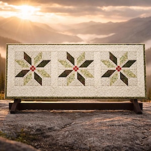 May include: A rectangular quilt with three starburst designs. Each star has green, brown, and red accents. The quilt is displayed on a wooden stand, set against a backdrop of mountains and a sunset.