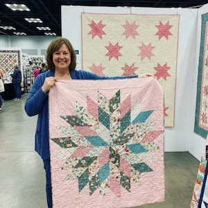 May include: A woman holding a handmade quilt with a starburst design. The quilt features a light pink background with alternating sections of floral fabric, teal, and pink. Another quilt with pink star patterns hangs in the background.