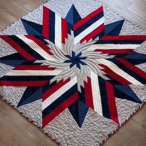 May include: A quilted star-shaped design in red, white, and blue. The center features a spiral pattern, with the star points radiating outwards. The quilt has a light beige background with a patterned border.