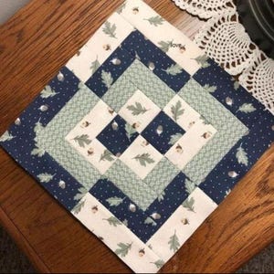May include: A square quilted placemat with a diamond pattern. The design features alternating bands of navy blue with white dots, light green with leaf patterns, and off-white with acorns and leaves. The word "love" is visible in the corner.