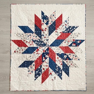 May include: A square quilt featuring a central star pattern composed of diamond-shaped fabric pieces in red, navy blue, and white floral prints. The background is a solid off-white fabric with intricate, swirling machine-stitched quilting patterns.
