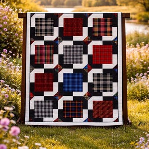 Op de afbeelding: Een kleurrijke quilt tentoongesteld op een houten standaard buiten. De quilt heeft een patchwork-ontwerp met vierkanten in rood, blauw, zwart en ruitpatronen, omlijst door witte en zwarte randen. De scène speelt zich af in een grasveld met bloemen en een waterpartij op de achtergrond.