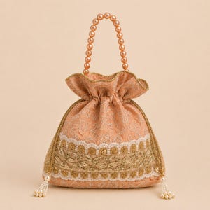 May include: A peach-colored drawstring pouch bag featuring intricate white floral embroidery, a wide band of gold metallic trim with scalloped edges, and a handle made of a single strand of round, glossy, light-colored faux pearls.