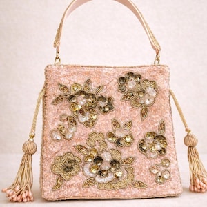 May include: A blush pink handbag with a top handle and floral embroidery. The bag is embellished with gold beads, sequins, and tassels. The handle and tassels are a matching light pink color.