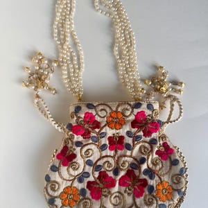 May include: An off-white embroidered bag with a pearl beaded strap. The bag features a floral design with red and orange flowers, blue leaves, and gold embroidery. The strap is made of multiple strands of small pearls.