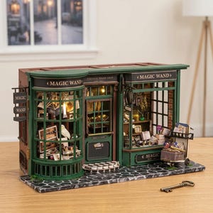 May include: Miniature model of a magic wand shop with green and brown exterior. The shop features detailed windows, shelves filled with items, and the text "MAGIC WAND". A key rests in front of the shop.