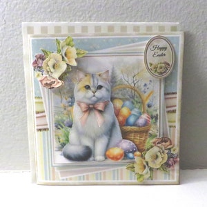 May include: A handmade Easter card featuring a white cat with a pink bow sitting next to a basket of colorful Easter eggs. The card has a blue background with a floral design and the text "Happy Easter" in a circle.