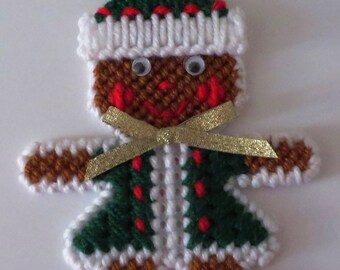 May include: A handmade gingerbread person Christmas ornament. The ornament is made of yarn and has a green and white striped shirt, a brown body, a white hat with red trim, and a gold ribbon. The ornament has googly eyes and a red mouth.