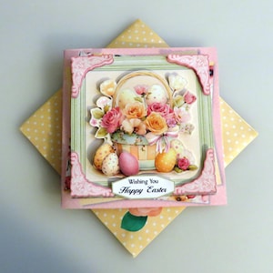 May include: A decorative Easter card featuring a basket of flowers and decorated eggs. The card has a pink frame with decorative corners and the text "Wishing You Happy Easter". The card is on a yellow polka dot background.