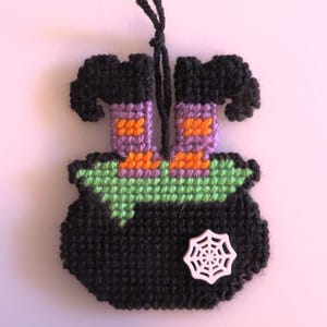 May include: A handmade Halloween decoration featuring a black cauldron with green and black trim, and a white spiderweb. The cauldron has a pair of witch legs in purple and orange. A black string is attached for hanging.