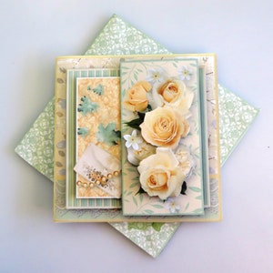 May include: A handmade card with a layered design featuring a bouquet of yellow roses with green leaves and white flowers. The card is decorated with a pearl-like string and has a green and white patterned background.