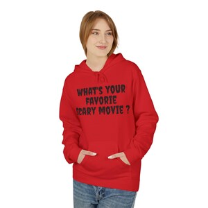 May include: A bright red hooded sweatshirt featuring a front kangaroo pocket and long sleeves. The chest displays the black text "WHAT'S YOUR FAVORIE SCARY MOVIE ?" in a jagged, horror-inspired font style. The garment is paired with blue denim jeans.