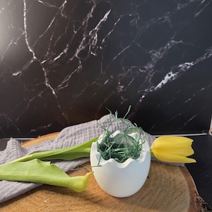 Könnte beinhalten: Ein weißer, eiförmiger Keramik-Pflanztopf, gefüllt mit grünem Ziergras, auf einer Holzoberfläche. Eine gelbe Tulpe und ein graues Tuch sind ebenfalls vorhanden. Der Hintergrund ist eine schwarz marmorierte Oberfläche.