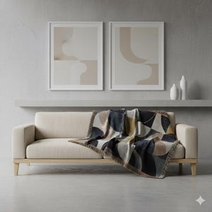 Puede incluir: Un sofá beige con patas de madera clara, adornado con una manta estampada geométrica en azul marino, beige y dorado. Dos impresiones artísticas abstractas enmarcadas cuelgan sobre el sofá, con jarrones blancos en un estante.