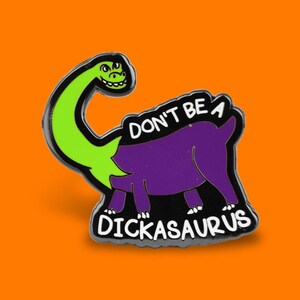 May include: A novelty enamel pin depicting a cartoon dinosaur with a green head and neck, a purple body, and the text "DON'T BE A DICKASAURUS" in white. The pin is set against an orange background.