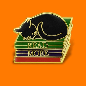 Pode incluir: Um alfinete dourado com um gato preto enrolado em uma pilha de livros. Os livros são verdes, roxos e vermelhos, com as palavras "READ MORE" em letras douradas. O alfinete tem um acabamento brilhante sobre um fundo laranja.