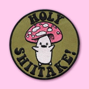 May include: Embroidered patch with a cartoon mushroom character with a smiling face and arms outstretched. The mushroom has a pink cap with white spots and the text "HOLY SHIITAKE!" in black lettering on a green background.