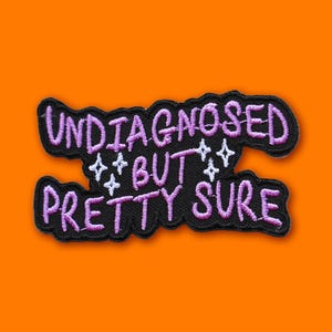 Peut inclure: Un écusson brodé noir avec les mots "UNDIAGNOSED BUT PRETTY SURE" en fil violet. L'écusson a une bordure noire et quatre accents blancs en forme d'étoile. Le fond est orange.